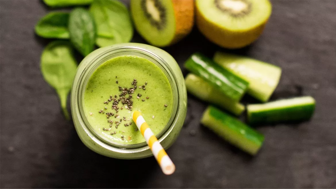 Photograph of a green smoothy in a mason jar with a red and white straw, surrounded by cucumber, kiwi, spinach on a slate tile. Shot by Paris Penny Photographer