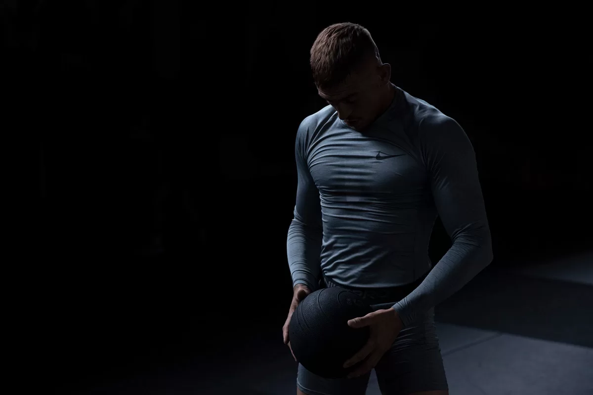 Dark contrasty photograph of a man in grey Nike gym wear training with a medicine ball in a dark moody gym. Photographed by Bristol Photographer Paris Penny