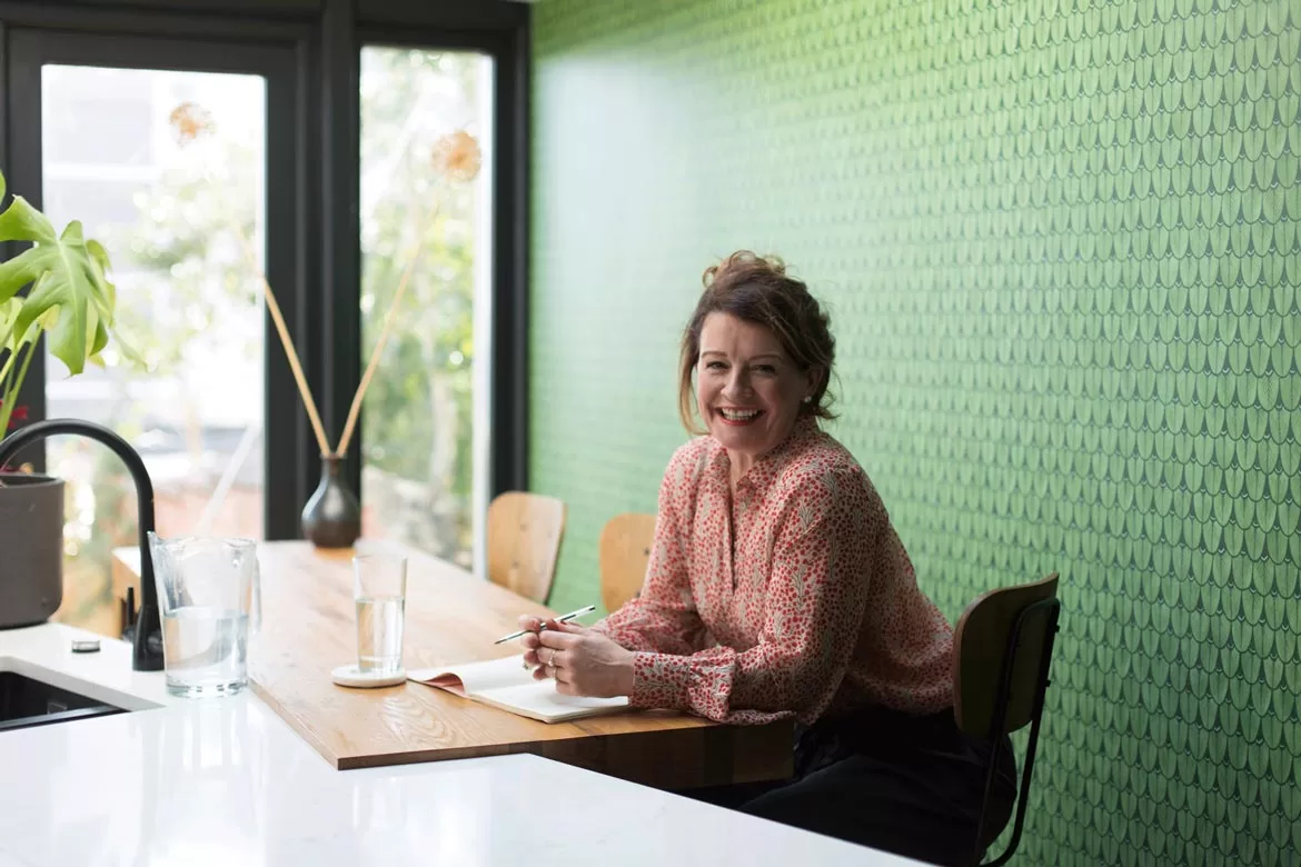 Photograph of a happy smiling woman sat at her stylish kitchen counter, with French door and green pattern wall paper.
